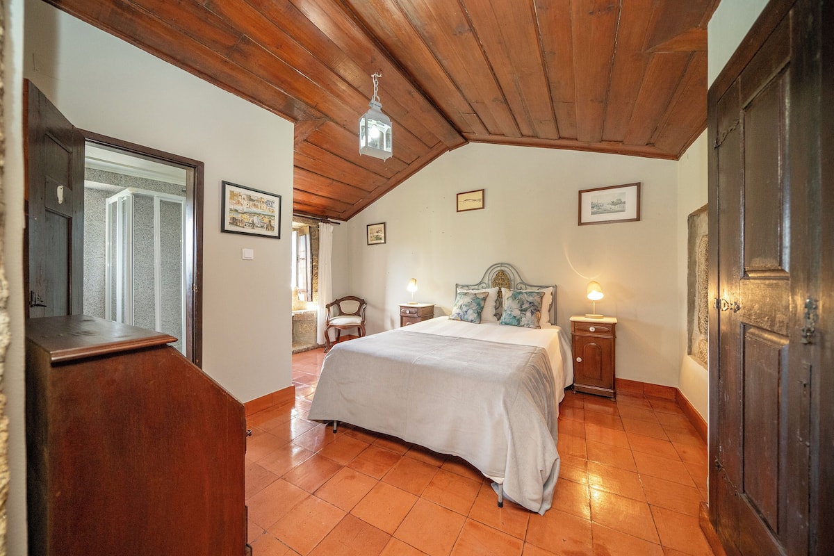 A bedroom with a wooden ceiling, a bed with a white bedspread, two bedside tables with lamps, and a wooden door.