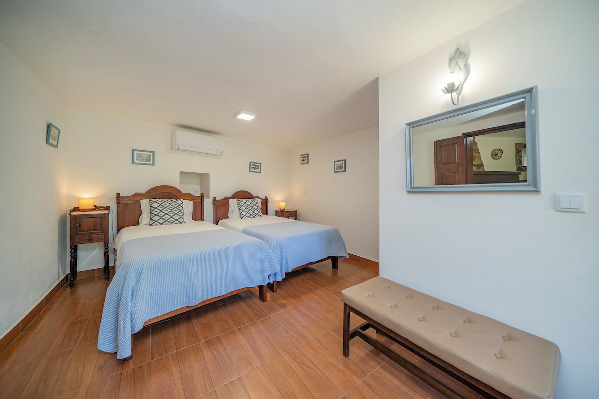 A hotel room with two beds, a bench, a wall-mounted mirror, and a bedside table with a lamp.