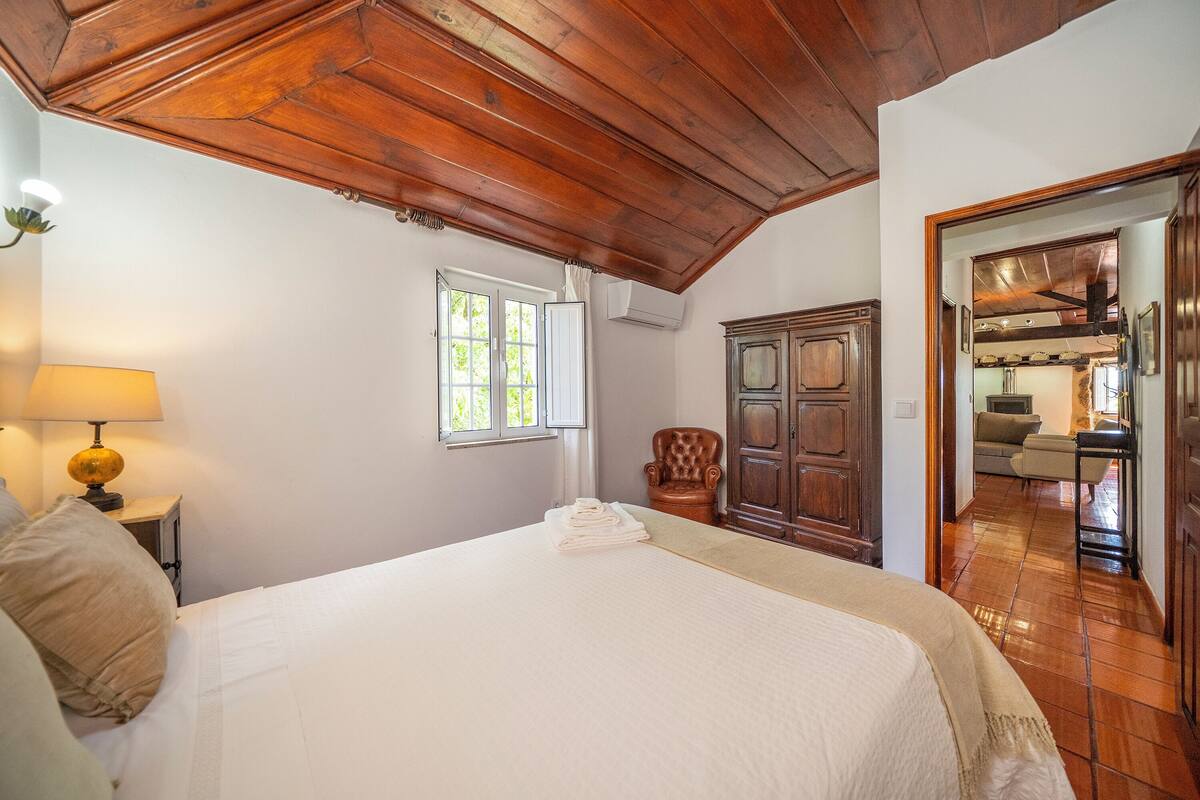 A bedroom with a wooden ceiling, a bed with white linens, a nightstand with a lamp, a window with curtains, and a wooden wardrobe.