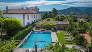 Una amplia piscina rectangular con solárium de piedra, rodeada de abundante vegetación y un edificio blanco con tejado de tejas rojas.