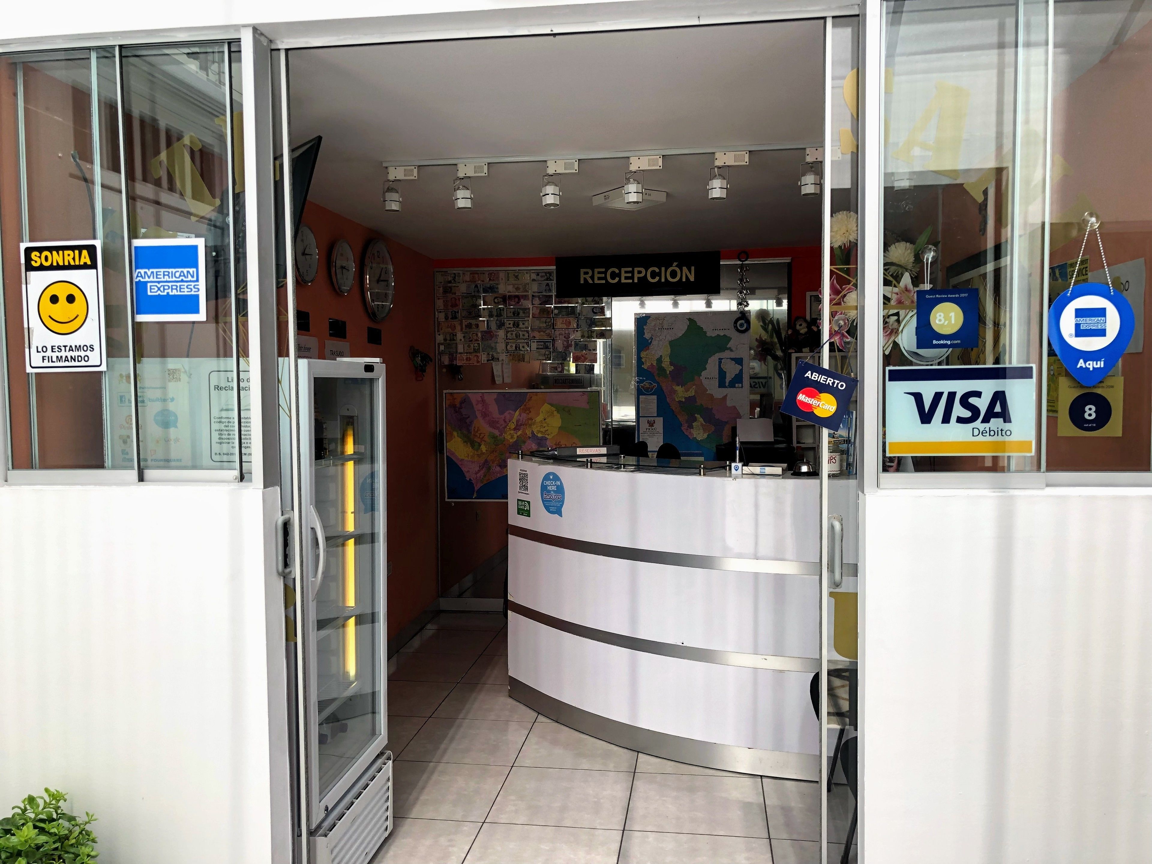 Un área de recepción con mostrador, carteles indicando que se aceptan Visa y MasterCard, un mapa en la pared y un letrero que indica que la recepción está abierta.