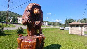 Una estatua de león de madera sobre un poste en un área con césped, con edificios y una carretera al fondo.