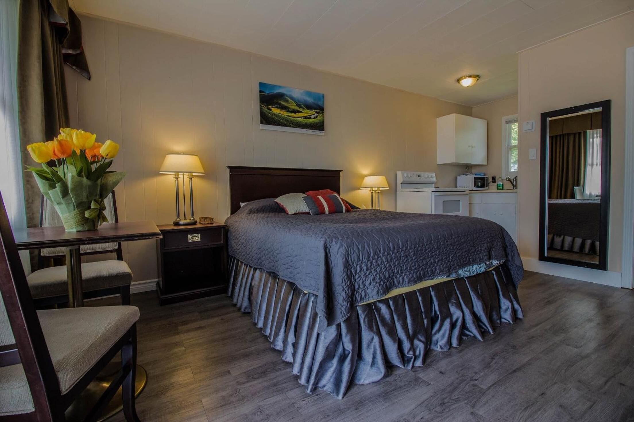 A bedroom with a bed, bedside tables, a desk, a chair, a lamp, a painting, and a kitchenette.
