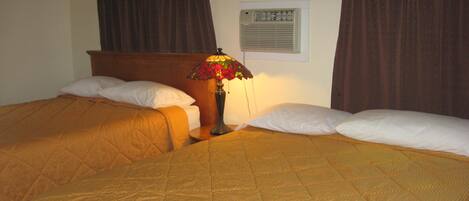 A hotel room with two beds, a nightstand, a lamp, and a wall-mounted air conditioning unit.