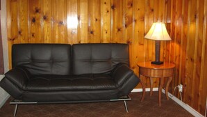 Ein schwarzes Ledersofa an einer holzverkleideten Wand, ein kleiner Holztisch mit einer Lampe und ein gemustertes Teppich.