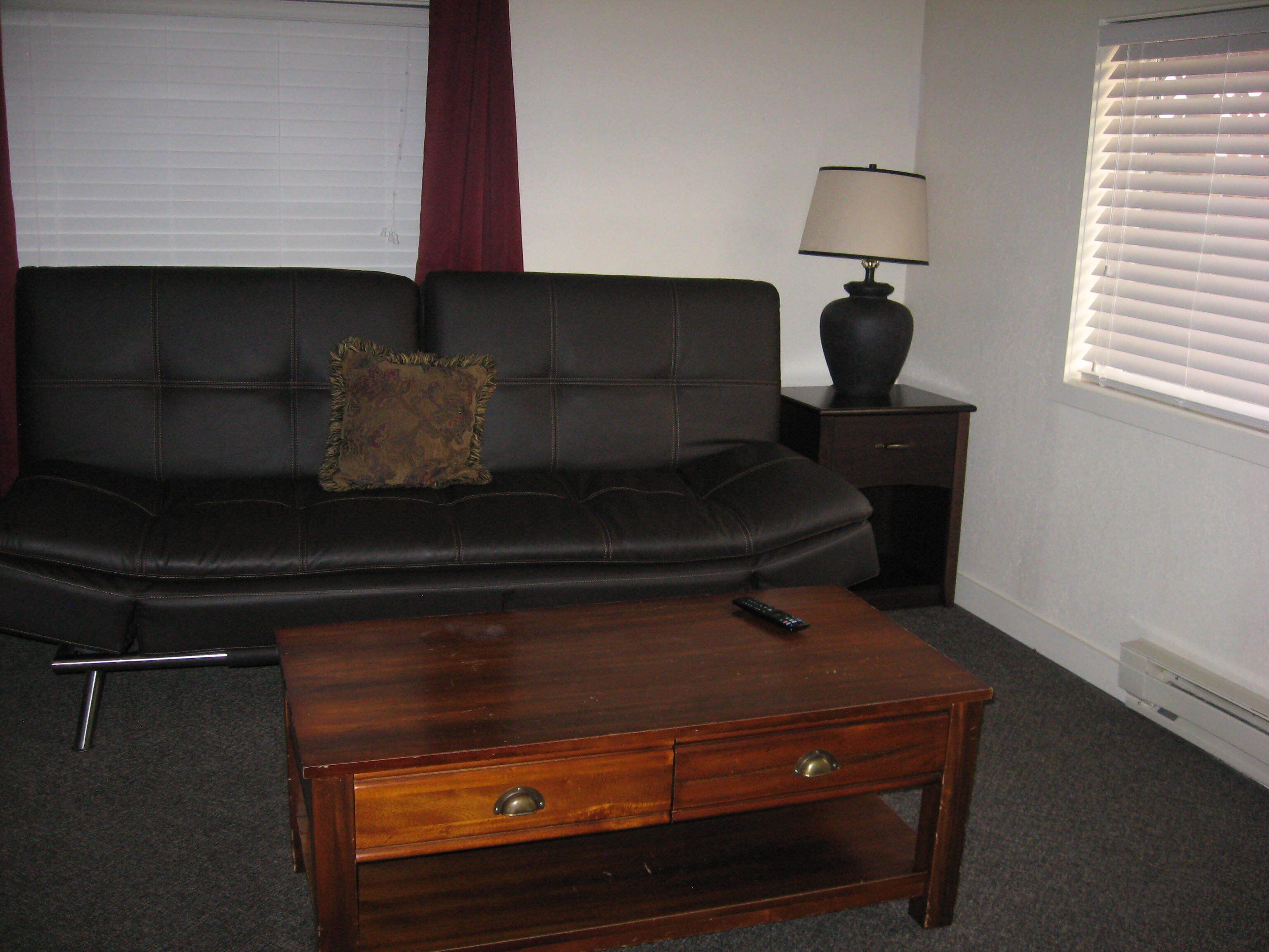 Una sala de estar con un sofá de cuero negro, una mesa de centro de madera con dos cajones, un control remoto sobre la mesa, una lámpara en una mesita auxiliar y una ventana con persianas.