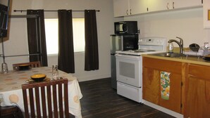 Eine kleine Küche mit Herd, Mikrowelle und Spüle. Ein Esstisch mit Stühlen und ein Fenster mit Vorhängen sind ebenfalls vorhanden.