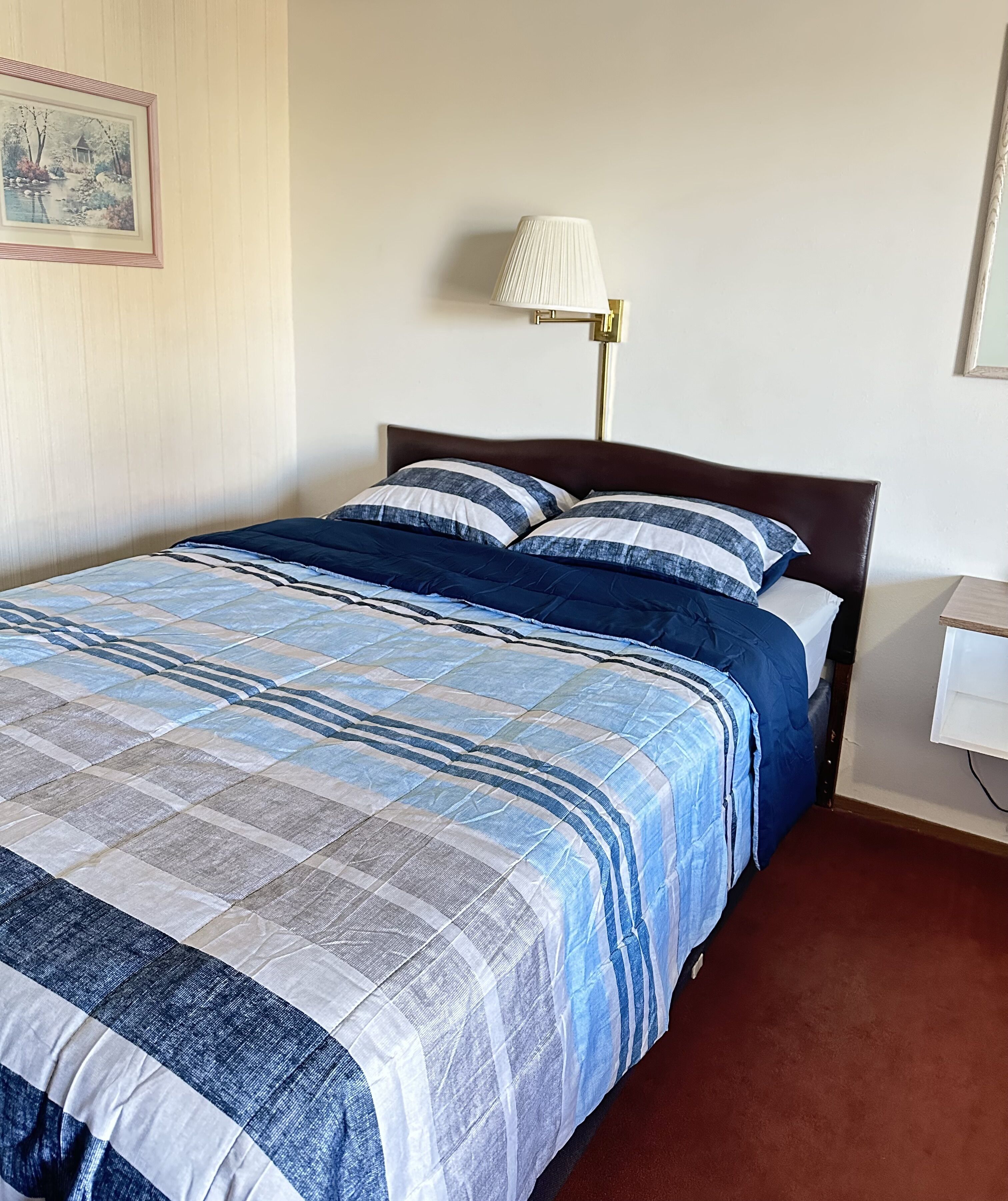 Una cama bien hecha con colcha azul y blanca a rayas, cabecera de madera oscura y lámpara de noche blanca.