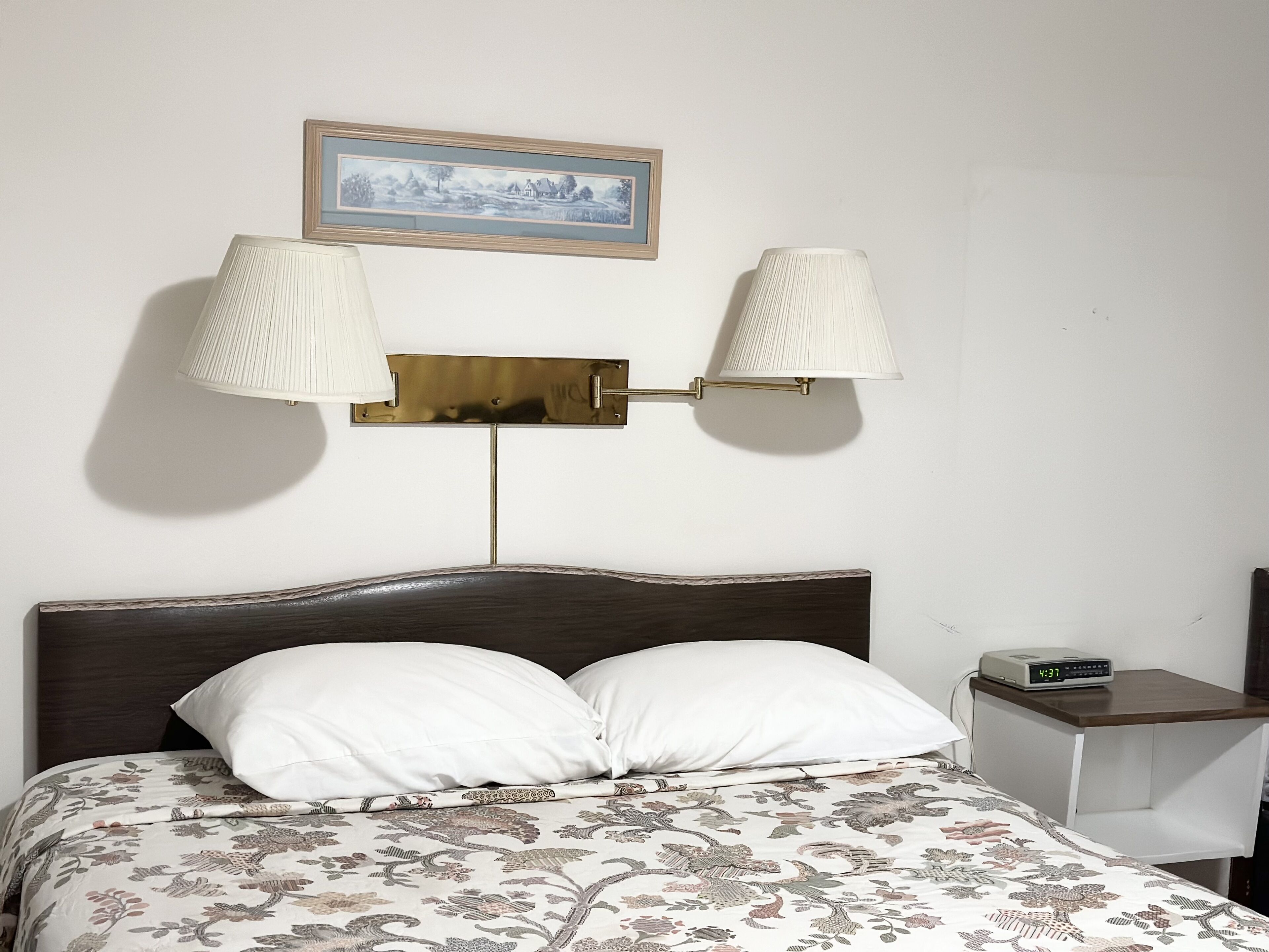A neatly made bed with a floral bedspread, two bedside lamps, a clock, and a framed picture on the wall.