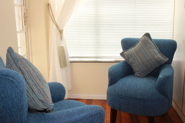 A room with a blue armchair and a matching ottoman, a window with blinds, and a curtain.