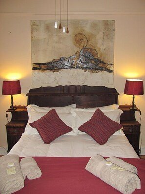 Una cama bien hecha con almohadones rojos, dos lámparas de noche y un cuadro abstracto grande sobre la cama.