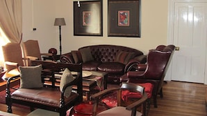 Un salon avec un canapé en cuir marron foncé, plusieurs chaises en bois, une table avec une lampe et deux tableaux encadrés accrochés au mur.