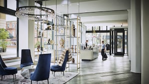 A modern hotel lobby with a reception desk, blue chairs, a chandelier, and a view of the street.