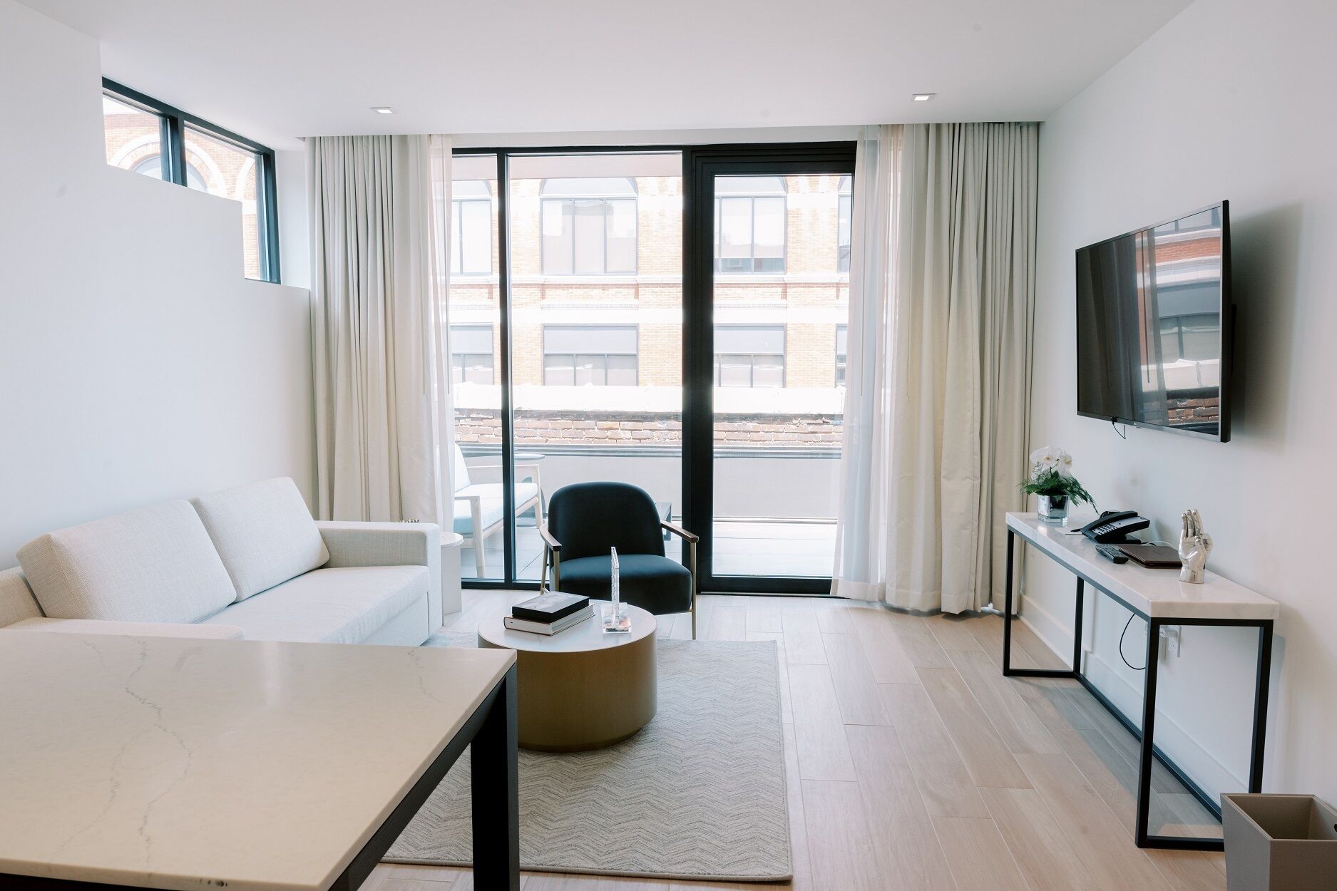 A modern hotel room with a sofa, a round table, a chair, a desk with a TV, and a view of the city through large windows.