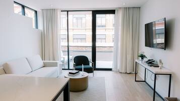 A modern hotel room with a sofa, a round table, a chair, a desk with a TV, and a view of the city through large windows.