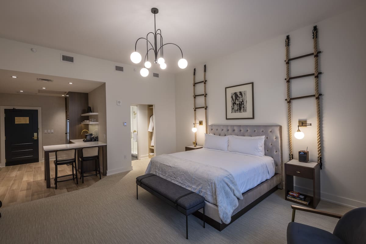 A modern hotel room with a large bed, a desk with two chairs, a bench, and a small table with a lamp.