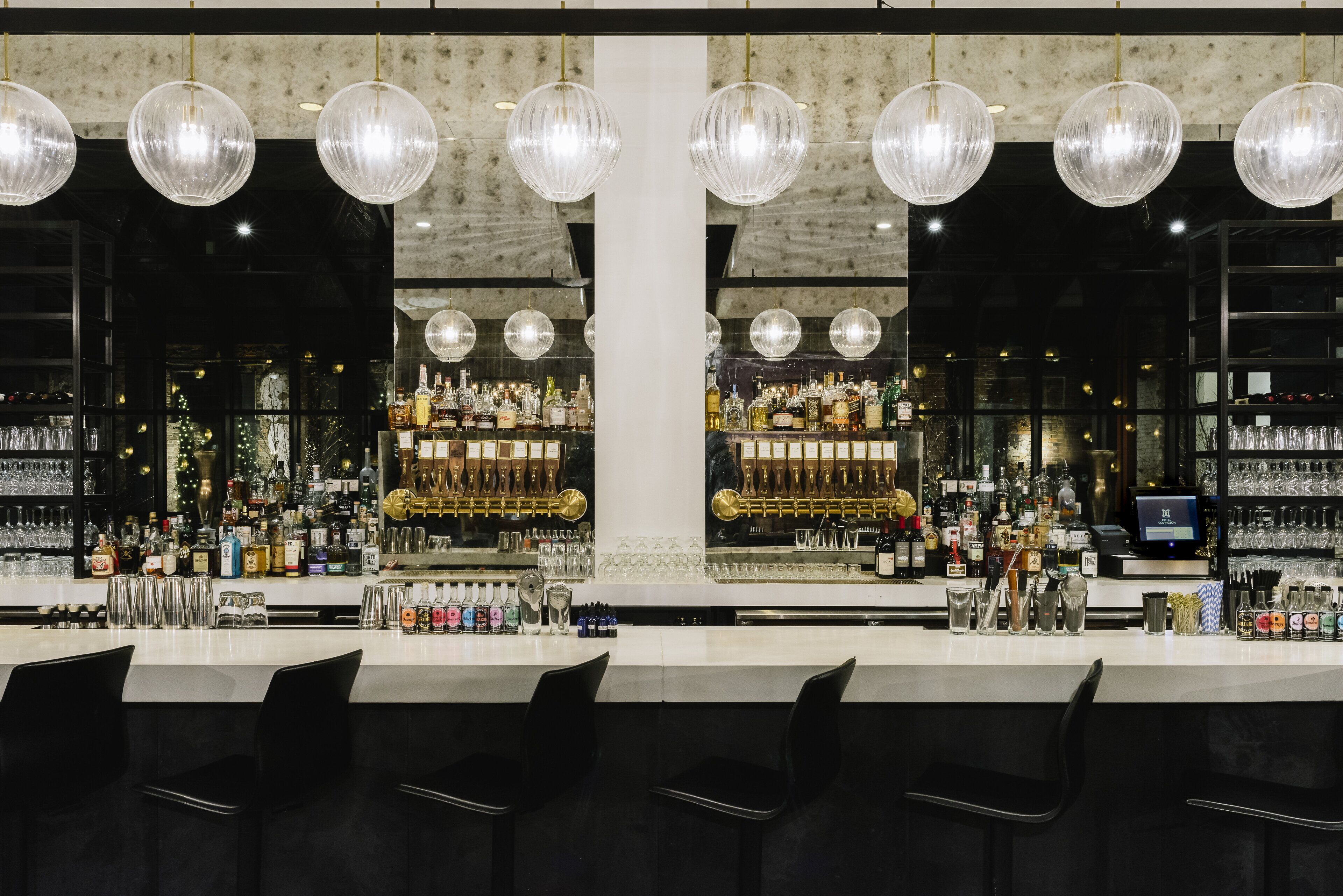 A bar with multiple hanging crystal chandeliers, a well-stocked bar counter, and black bar stools.