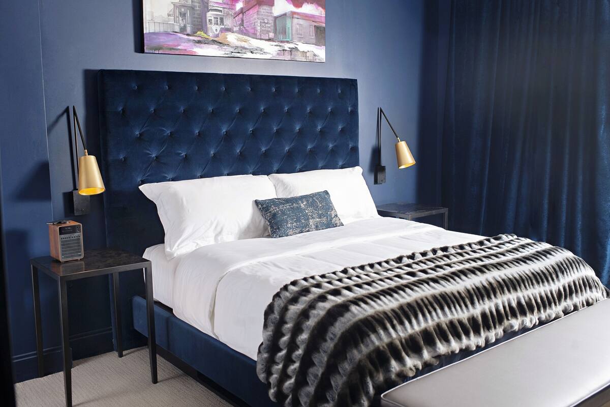 A bedroom with a tufted headboard, white bedding, a black and white throw blanket, and two bedside lamps.