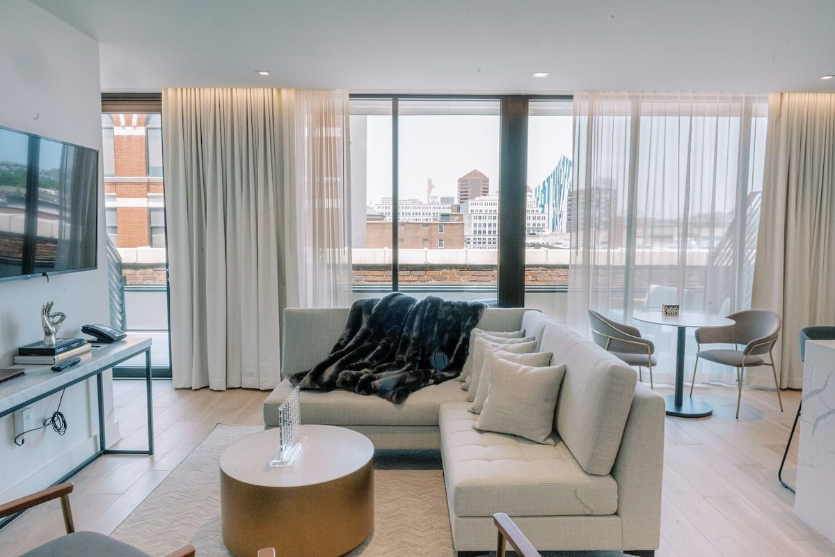 A modern living room with a sofa, a coffee table, and a TV. There is a dining area with a table and chairs. The room has large windows with curtains.