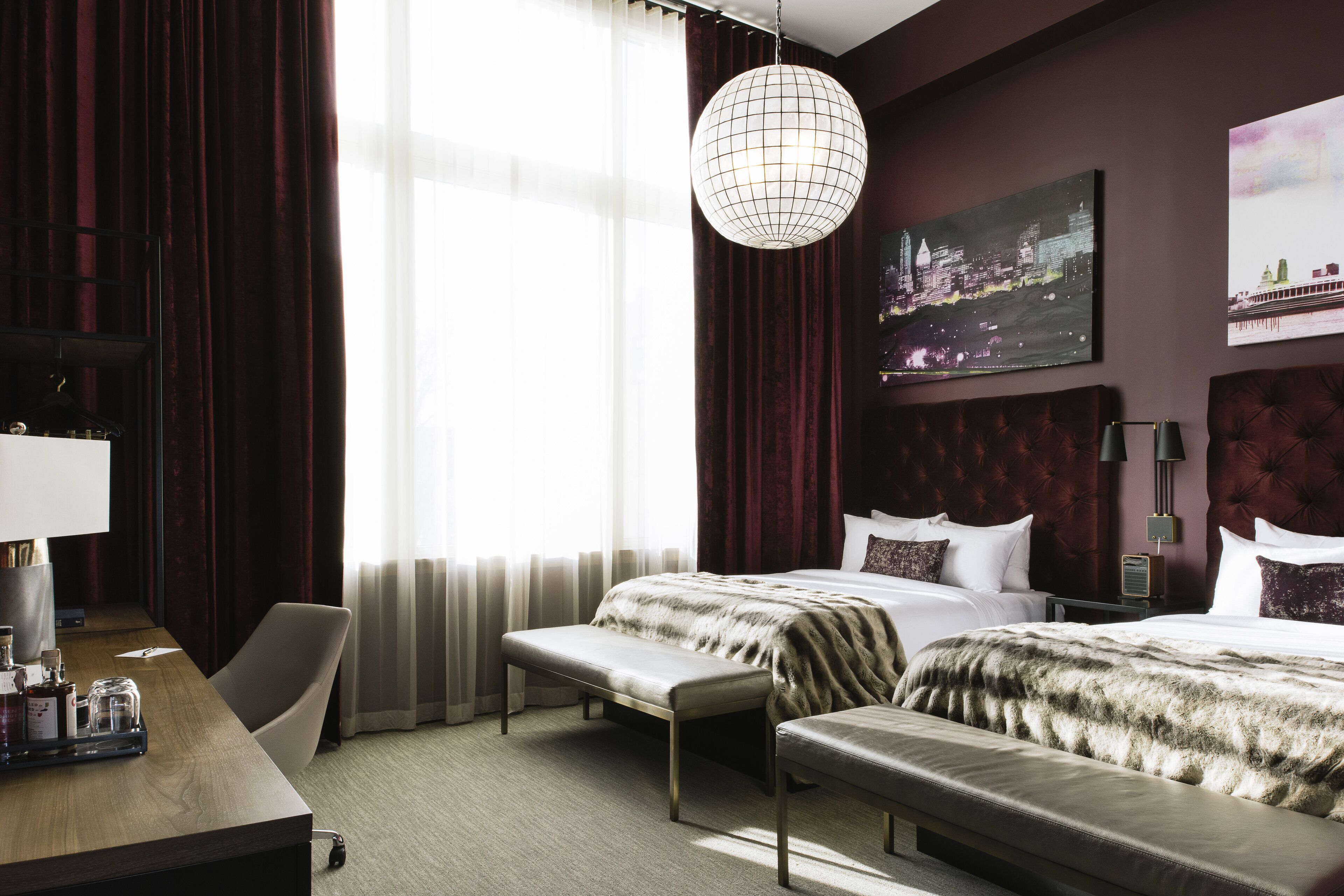 A hotel room with a large bed, a desk with a chair, a bench, a hanging light fixture, and a cityscape painting on the wall.