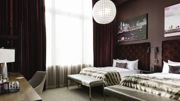 Habitación de hotel con una cama grande, un escritorio con silla, un banco, una lámpara colgante y un cuadro de un paisaje urbano en la pared.