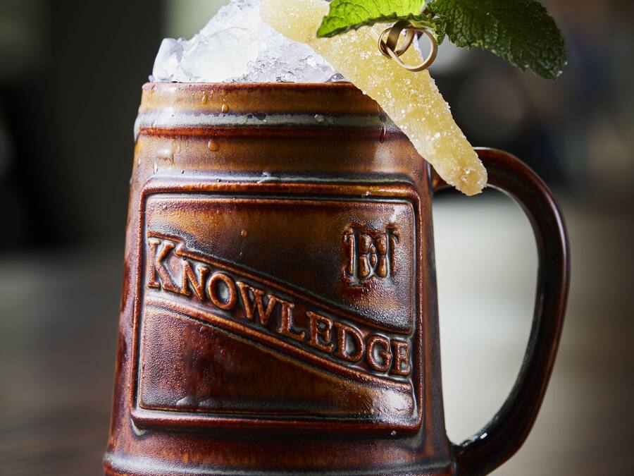 Eine hölzerne Tasse mit dem Prägezeichen 'KNOWLEDGE', gefüllt mit einem Getränk, Zucker und einem Minzblatt.