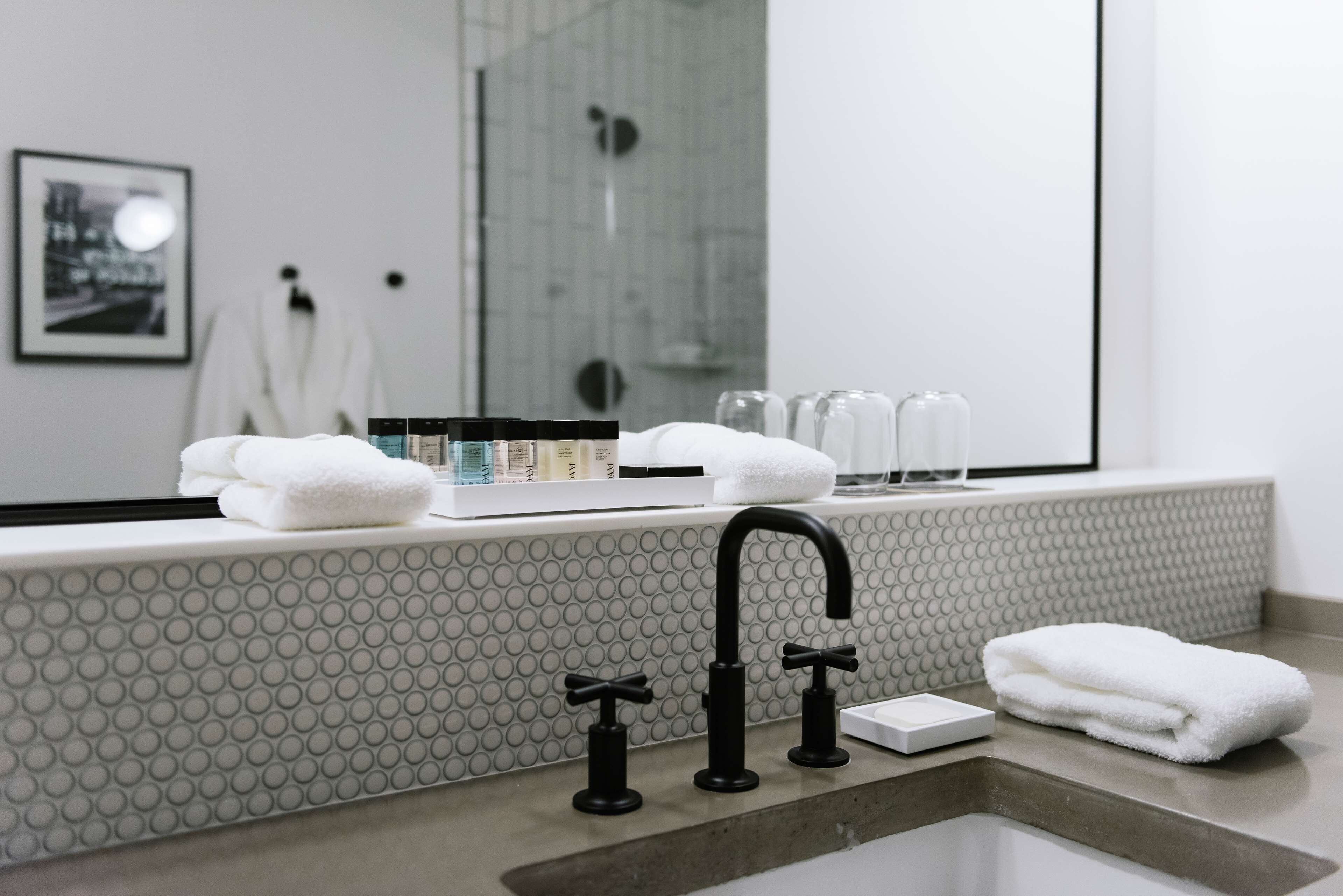 Ein Badezimmer im modernen Stil, mit einem großen Spiegel, einer Badewanne und einem Waschbecken mit zwei schwarzen Wasserhähnen. Auf der Ablage liegen Handtücher und Pflegeprodukte.