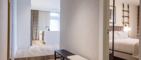 A modern hotel room with a wooden floor, a bench, a bed, and a view of the kitchen and living area.