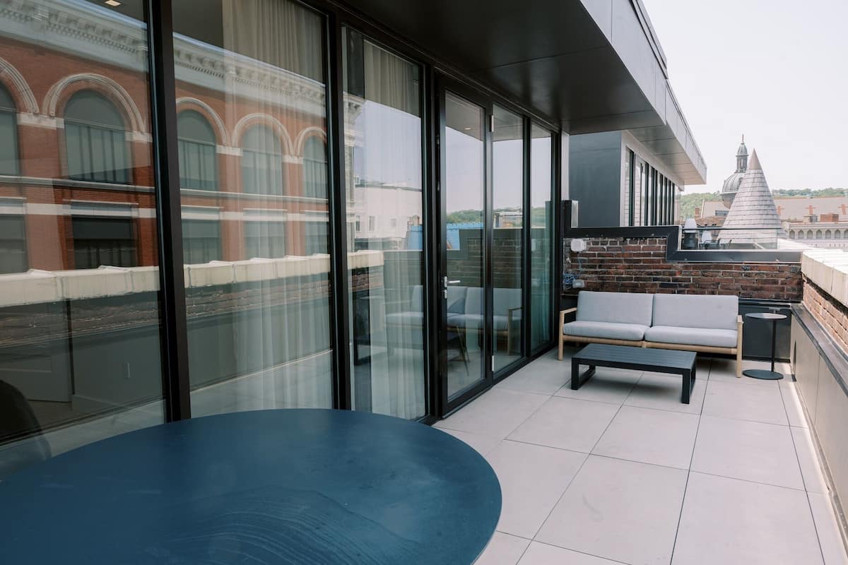 A modern outdoor seating area with a blue table and a grey sofa, large glass windows, and a view of a historic building in the background.