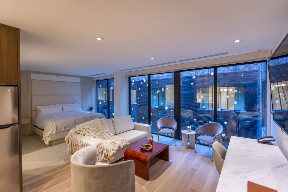 A modern hotel room with a bed, sofa, chairs, a small table, and large windows offering a view of the city.