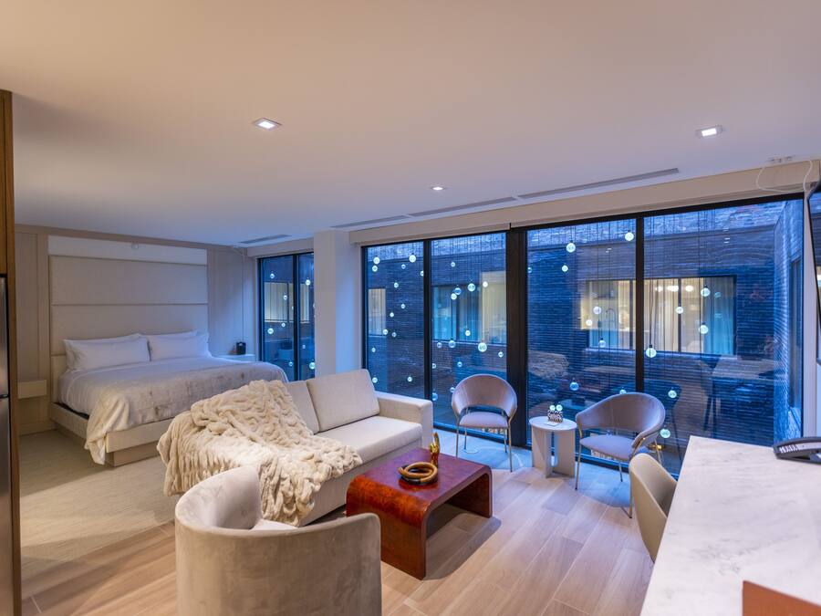 A modern hotel room with a bed, sofa, chairs, a small table, and large windows offering a view of the city.
