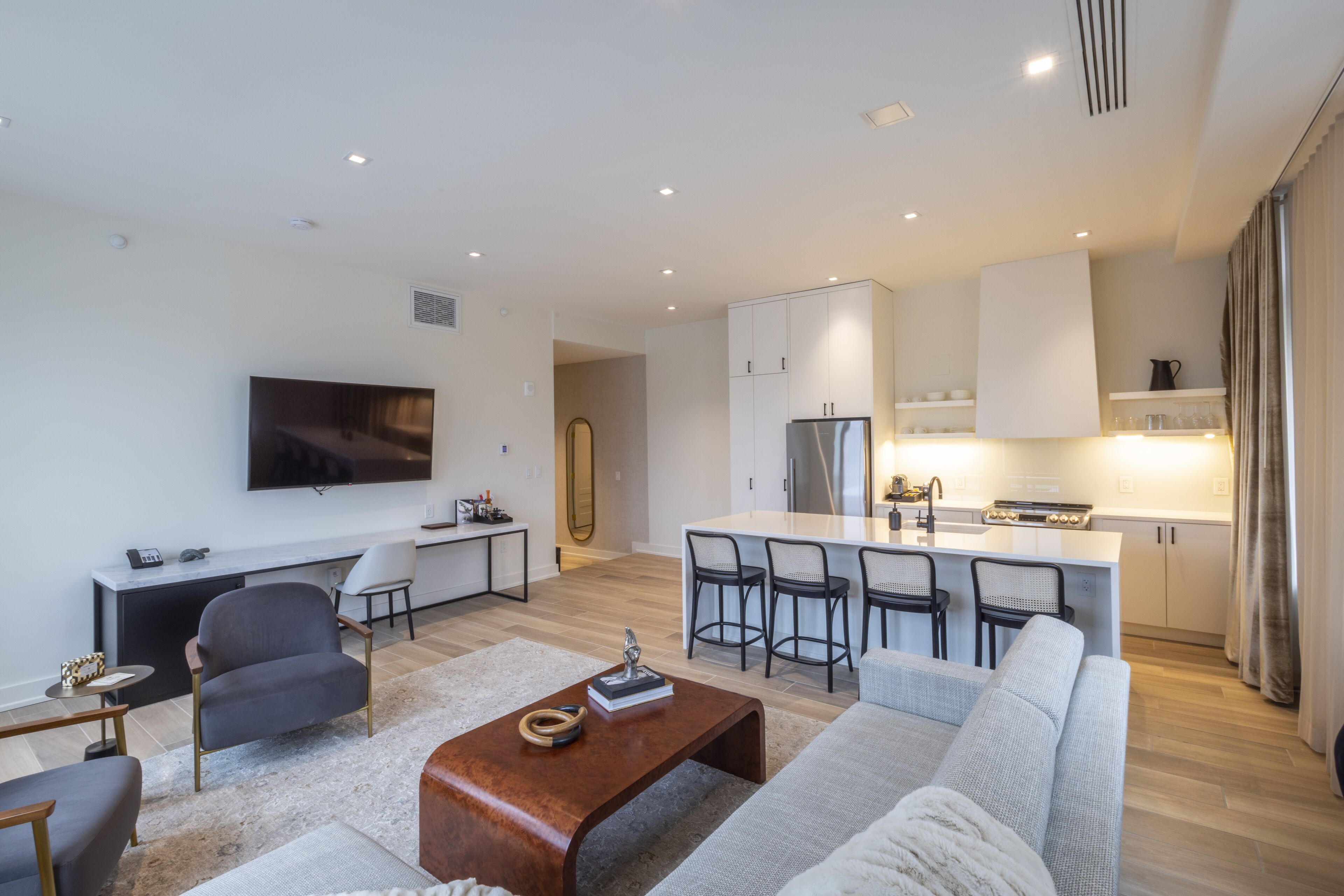 A modern living room with a sofa, coffee table, and TV. It includes a kitchen with a fridge, stove, and bar stools.