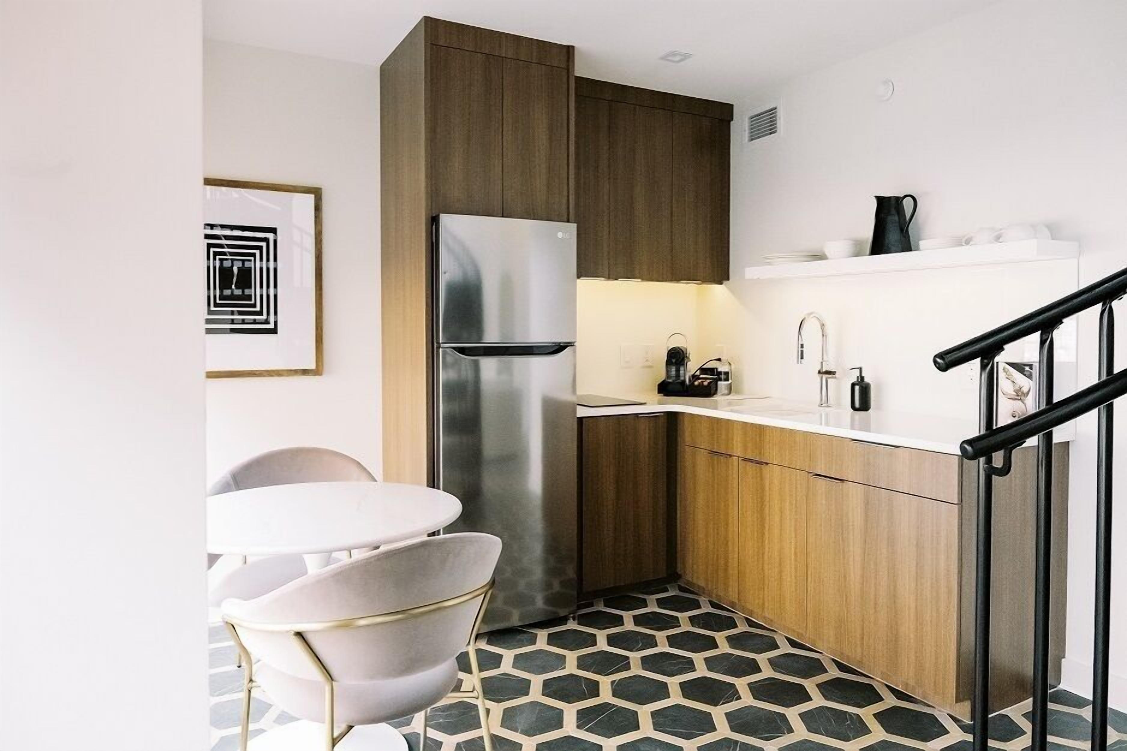 A compact kitchen with a stainless steel refrigerator, wooden cabinets, a white countertop, and a hexagonal patterned floor.