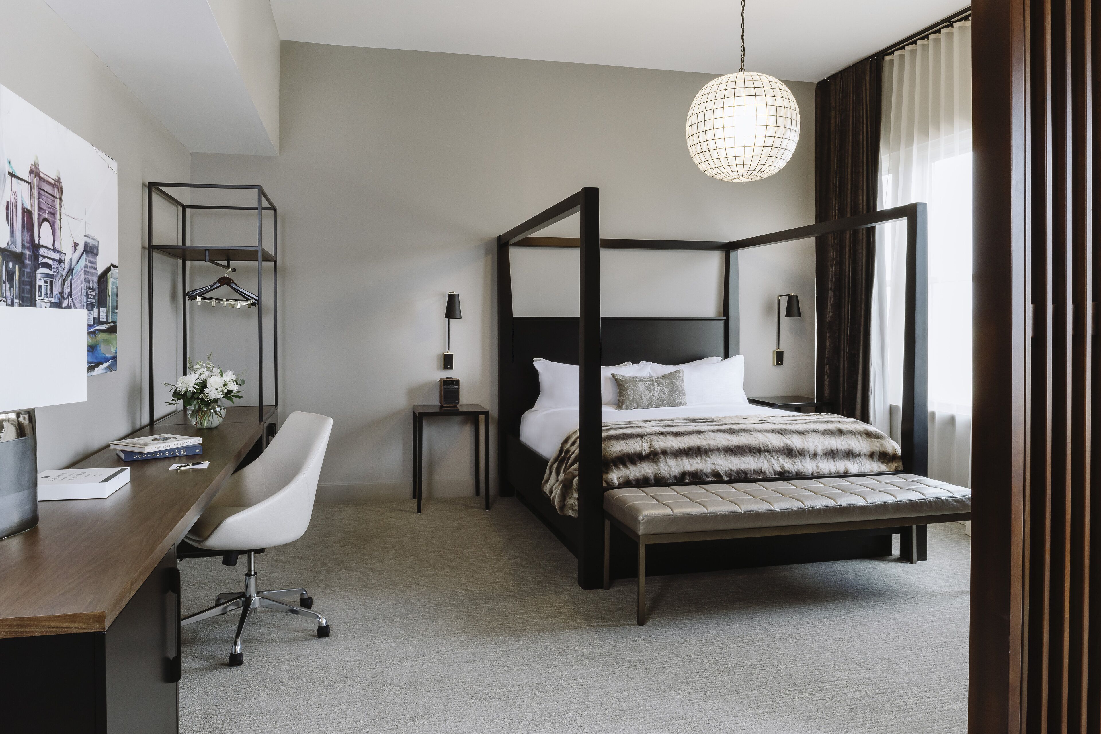 A modern hotel room with a four-poster bed, a desk with a chair, a bedside table with a lamp, and a large window with curtains.