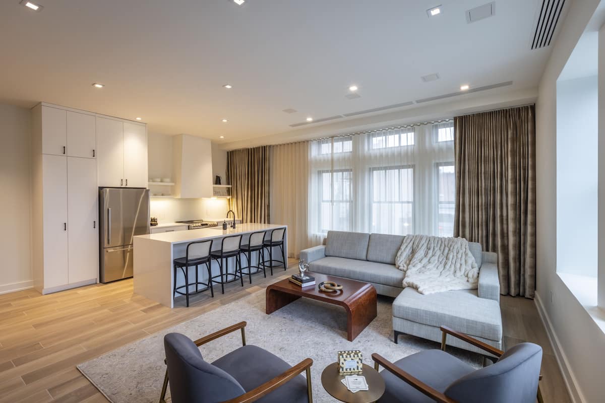 A modern living room with a sofa, two armchairs, a coffee table, and a kitchen area with a refrigerator and bar stools.