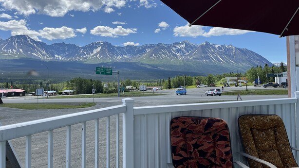 A view from a porch with a mountain backdrop, featuring a road sign, a parking area, and a few vehicles. - Kluane Park Inn (Haines Junction)