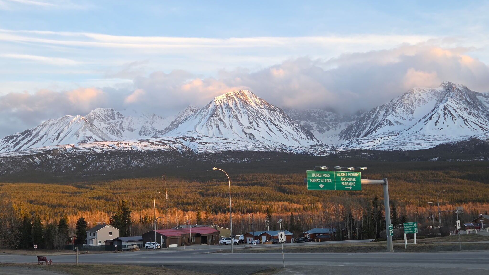 Wegwijzer met instructies naar Haines Road en de Alaska Highway, met een achtergrond van besneeuwde bergtoppen en een klein dorp met gebouwen en een weg.