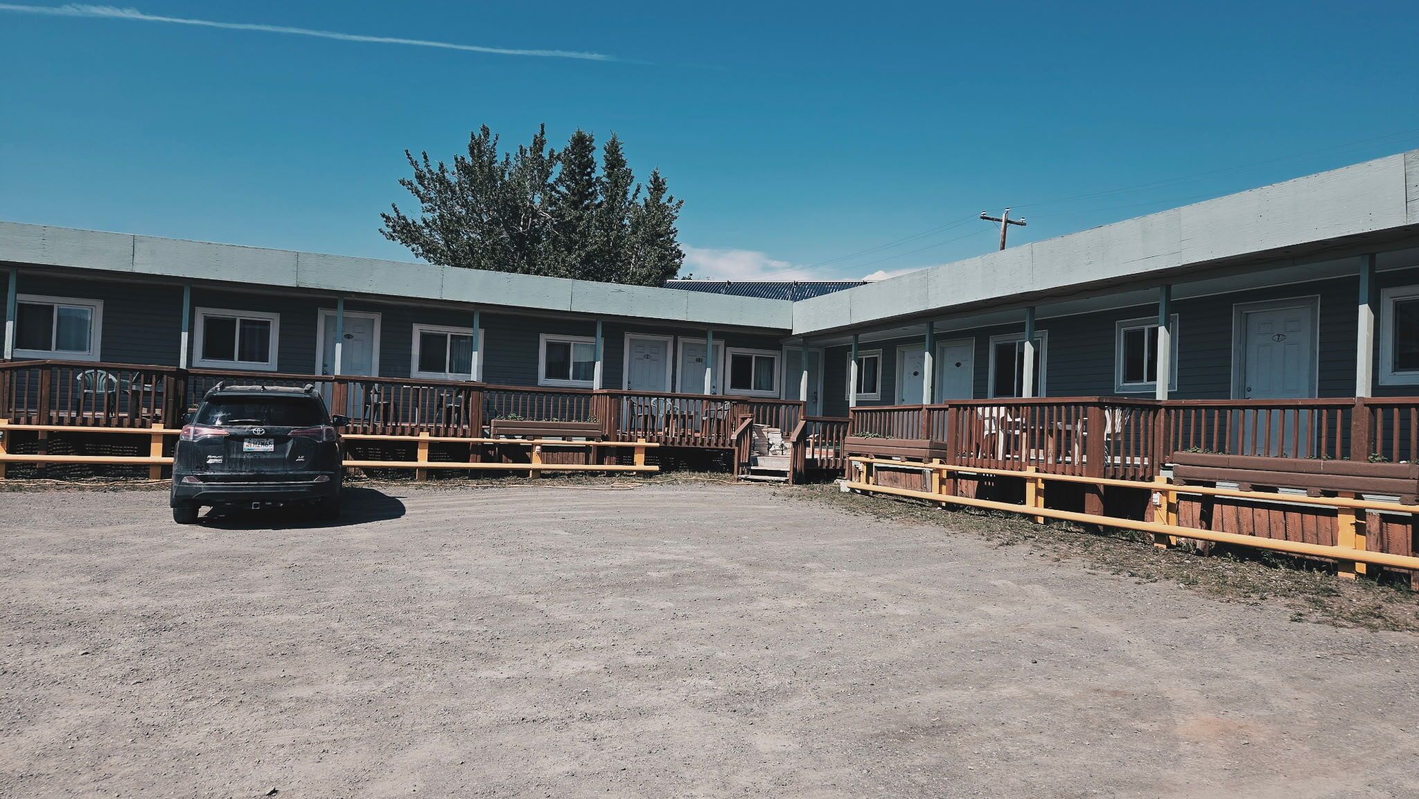 Een rij motelachtige gebouwen met individuele kamers, een parkeerplaats met grind en een zwart voertuig ervoor geparkeerd.
