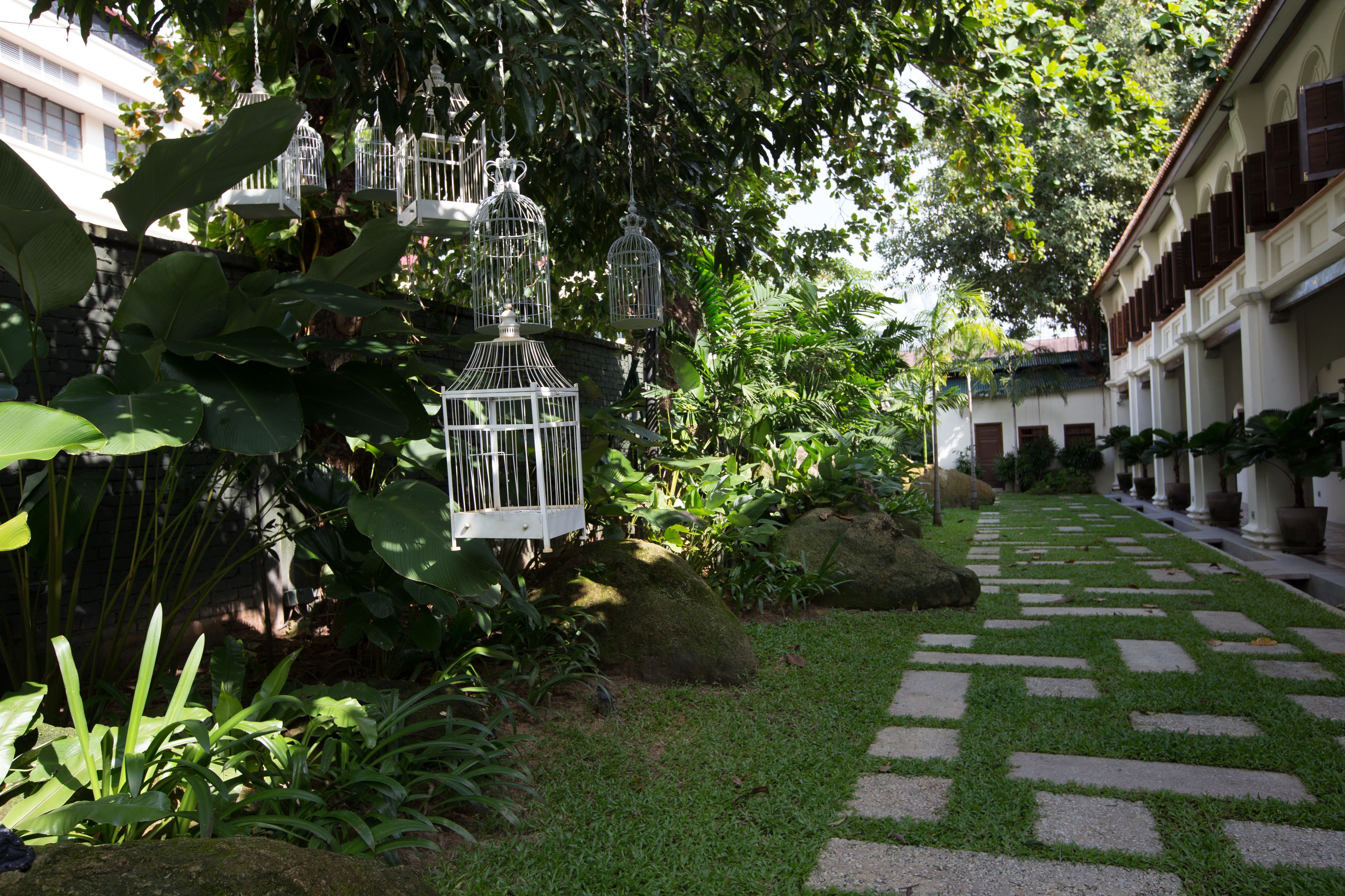A garden pathway with white birdcages hanging from trees, surrounded by lush greenery and a stone-tiled walkway.