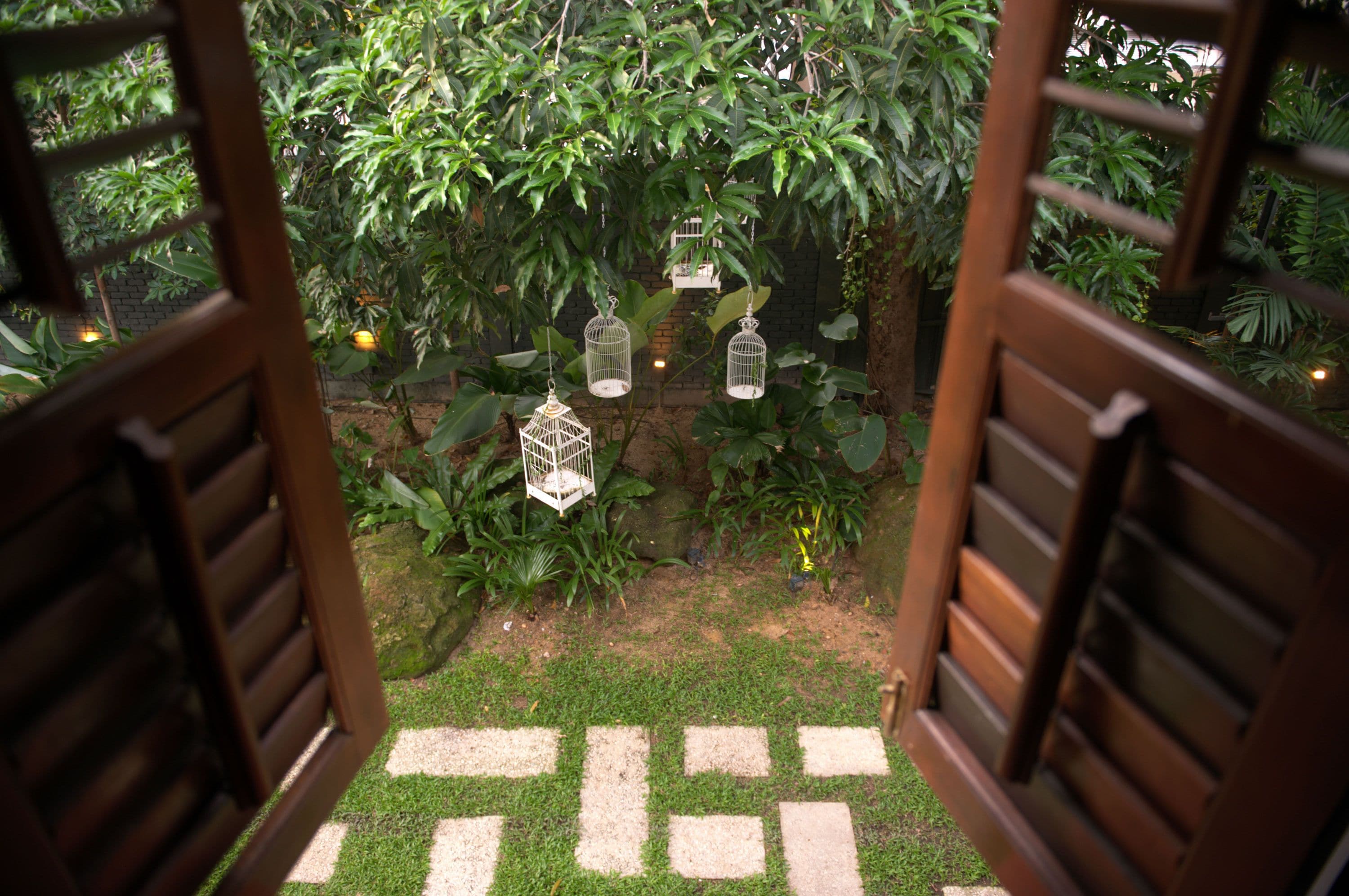 Un sendero en un jardín que conduce a una puerta de madera, rodeado de abundante vegetación y jaulas de pájaros colgantes.