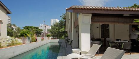 Piscina en la azotea con agua cristalina, rodeada de sillas de descanso y una zona de estar cubierta.