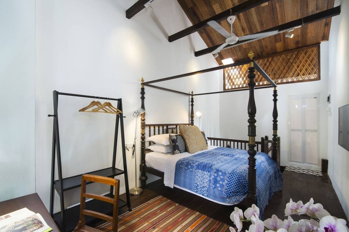 A four-poster bed with a blue patterned bedspread, a wooden chair, a striped rug, and a wooden ceiling.