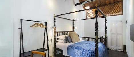 A four-poster bed with a blue patterned bedspread, a wooden chair, a striped rug, and a wooden ceiling.