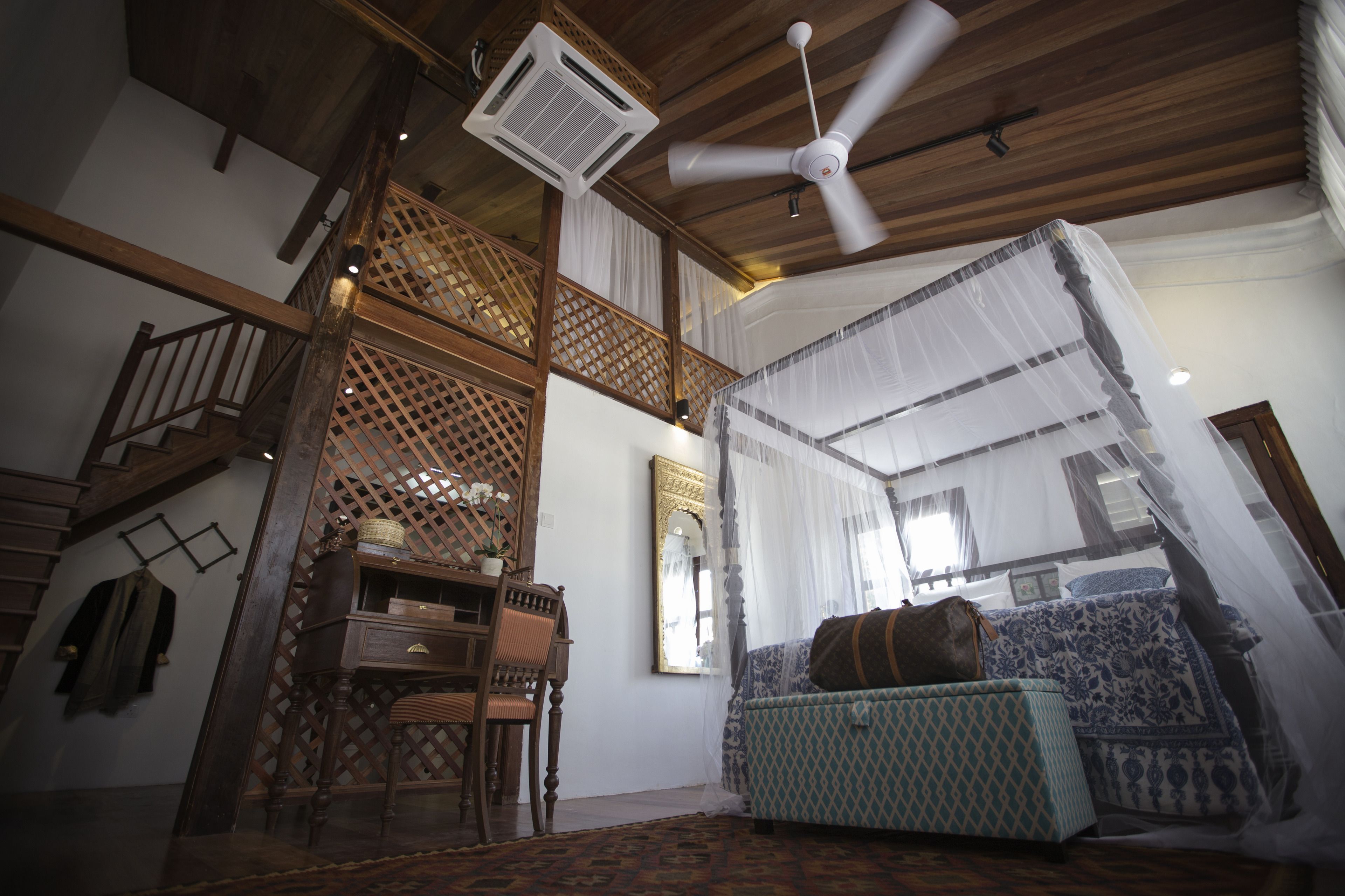 A room with a wooden ceiling, a ceiling fan, a bed with a canopy, a wooden chair, a small table, and a staircase.