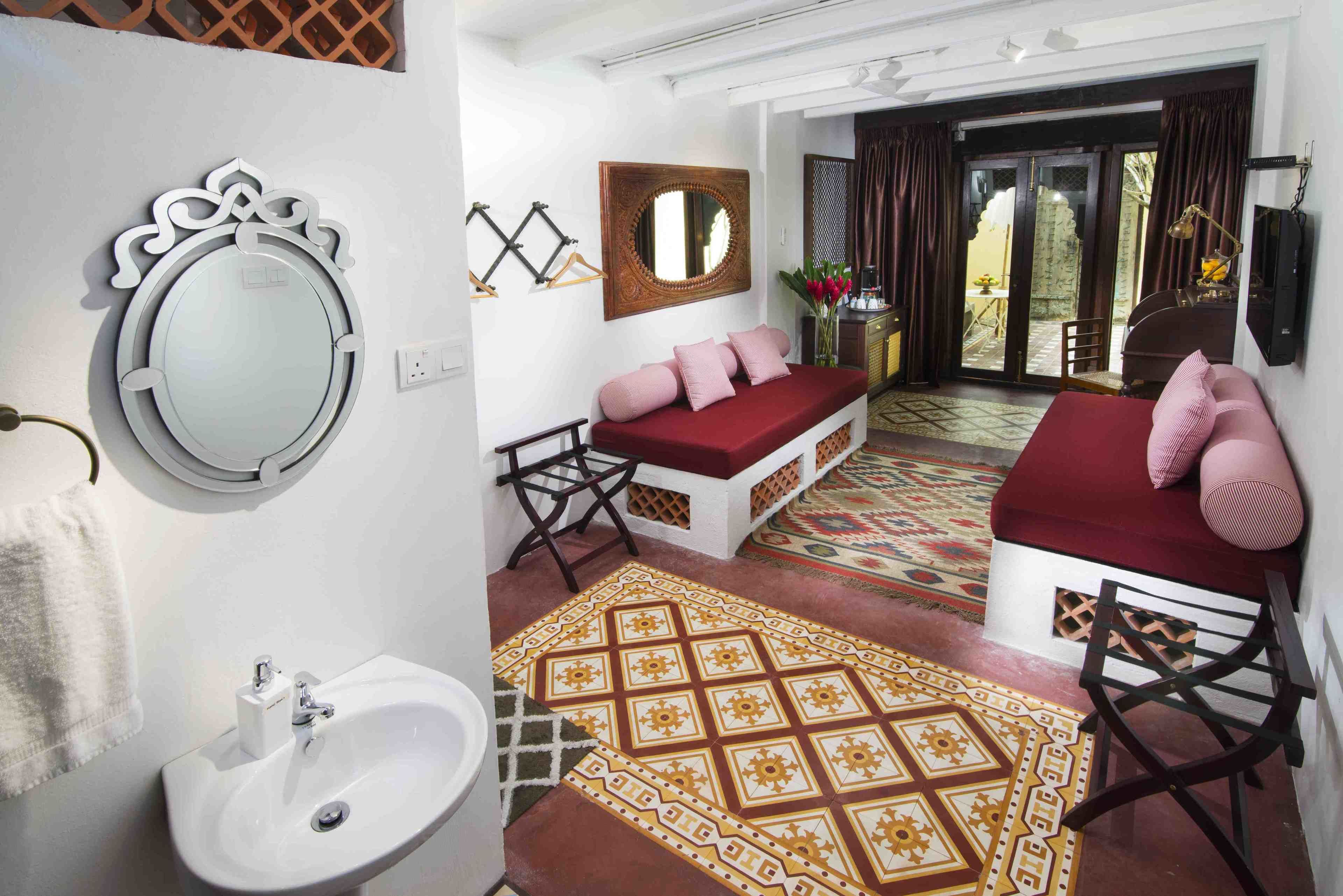 A hotel room with a white wall, a round mirror, a red sofa, a wooden chair, a small table with a lamp, and a patterned rug.