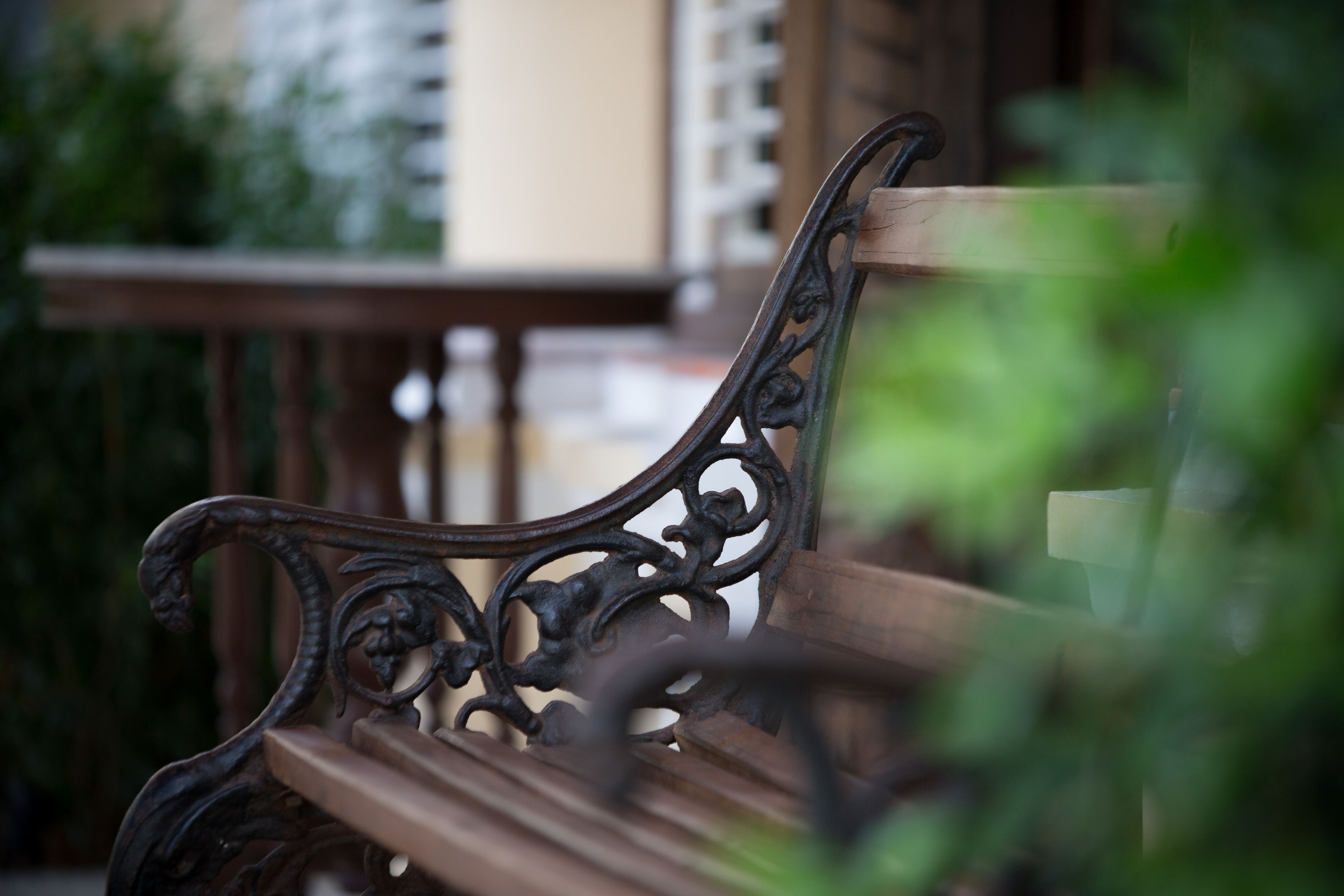 Un banc en bois orné de motifs métalliques, placé devant un arrière-plan flou de verdure et la façade d’un bâtiment.