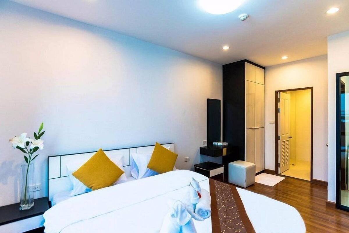 Habitación de hotel moderna con una cama bien hecha, una mesita de noche con un florero, un taburete pequeño y un vestidor.