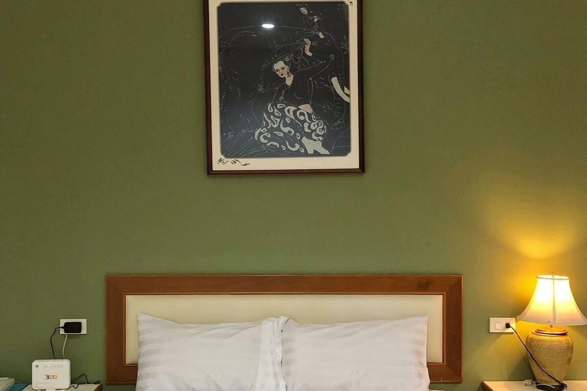 Ein Hotelzimmer mit Bett, zwei Kissen, einem Nachttisch mit Lampe, einem gerahmten Bild an der Wand und einem gefalteten Handtuch auf dem Bett.