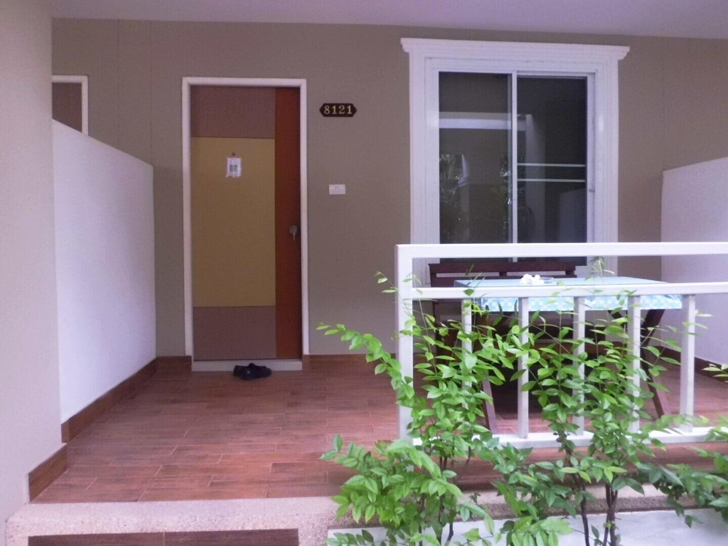 A small balcony with a table and chairs, a door with a number sign, and a window with blinds.