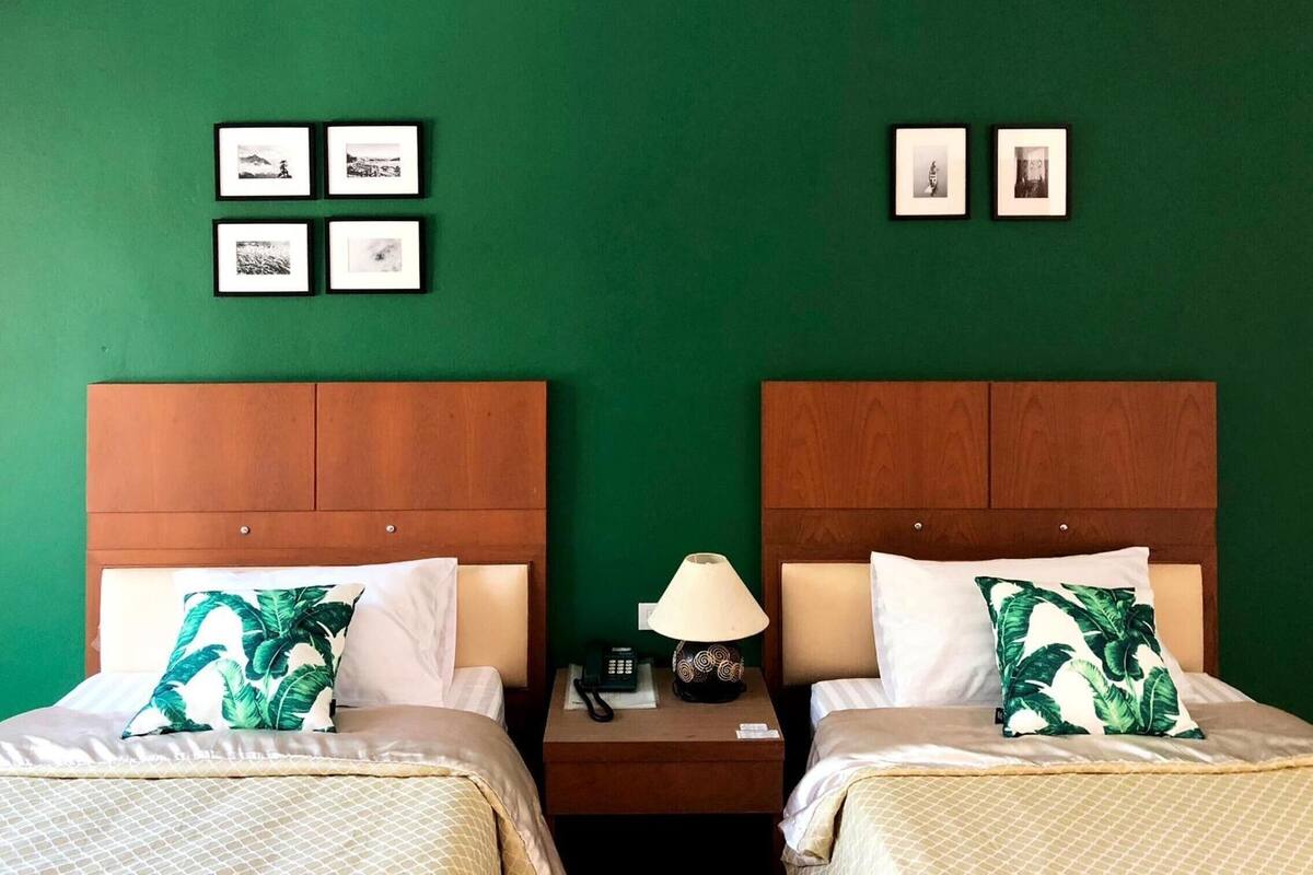 Ein Hotelzimmer mit zwei Betten, grünen Wänden, gerahmten Bildern an der Wand und einem Nachttisch mit Lampe.